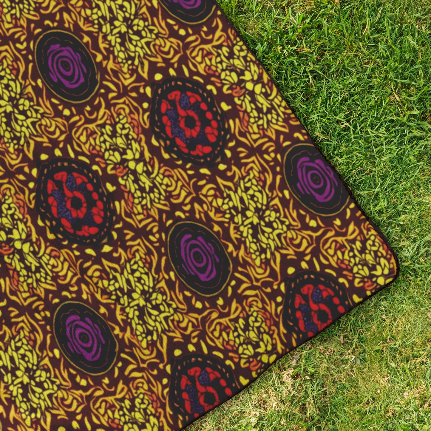 A colorful patterned fabric with swirling yellow, red, and purple designs lies on green grass. The visible corner showcases African-inspired patterns and intricate yellow shapes on a brown background, reminiscent of a plush fleece top.