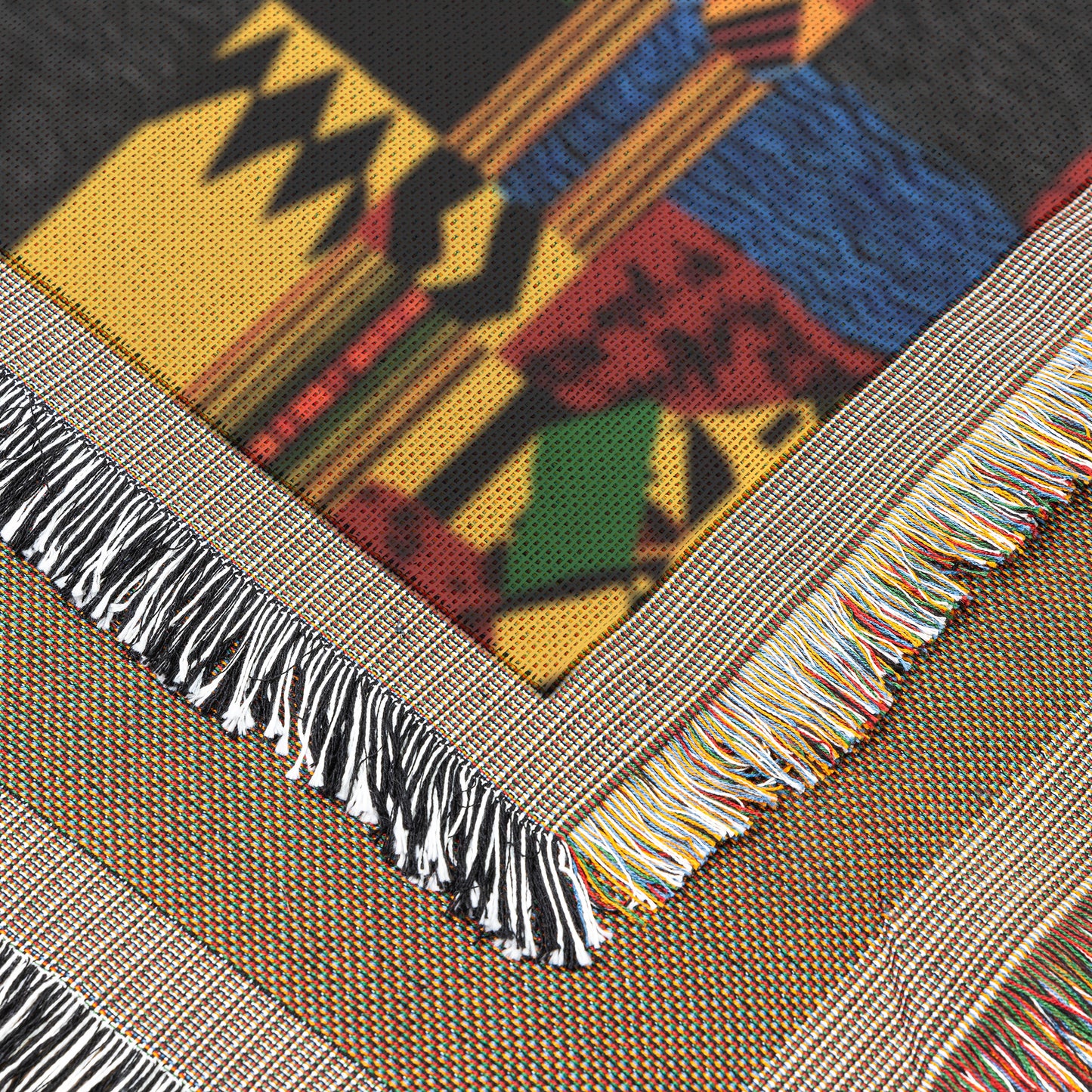 Close-up of a vibrant throw blanket with bold geometric patterns in yellow, black, red, blue, and green. Two fringed edges intersect at an angle, showcasing multicolored threads and intricate designs for a handcrafted look.