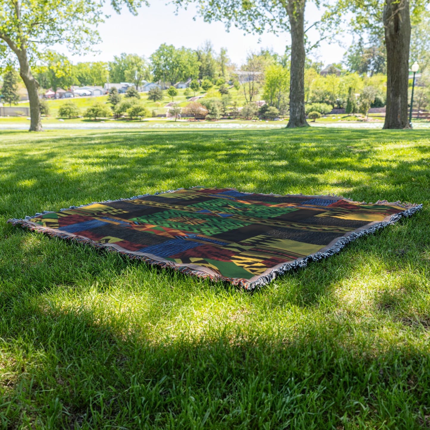 A cozy woven blanket with a colorful abstract pattern lies flat on green grass in a sunlit park. Trees with leafy branches cast dappled shadows, and a landscaped area with bushes and a walkway is visible in the background.