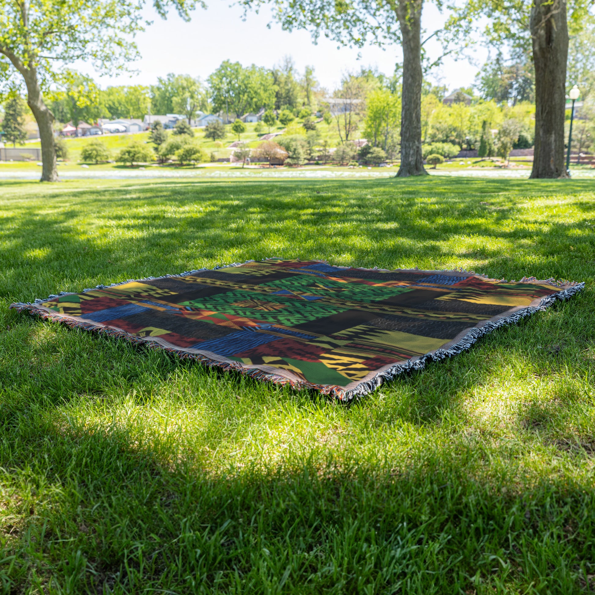 A cozy woven blanket with a colorful abstract pattern lies flat on green grass in a sunlit park. Trees with leafy branches cast dappled shadows, and a landscaped area with bushes and a walkway is visible in the background.