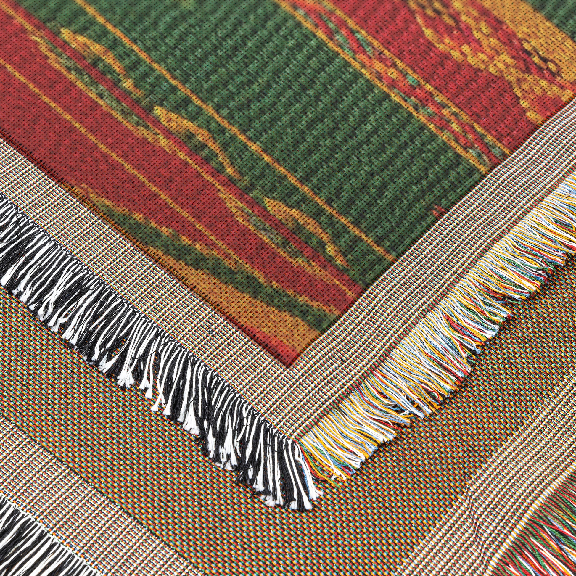 Close-up of two overlapping woven rugs with colorful fringe edges. The top rug showcases an African-inspired green, red, and yellow geometric pattern, while the rug underneath features a more subtle, earthy palette with multicolored fringed borders.