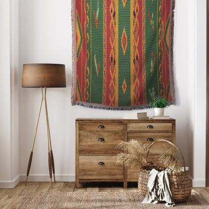 A cozy, African-inspired room features a wooden dresser, a basket with dried plants, a tall tripod floor lamp, and a colorful geometric woven tapestry with green, red, and yellow patterns. White walls and a wooden floor complete the space.