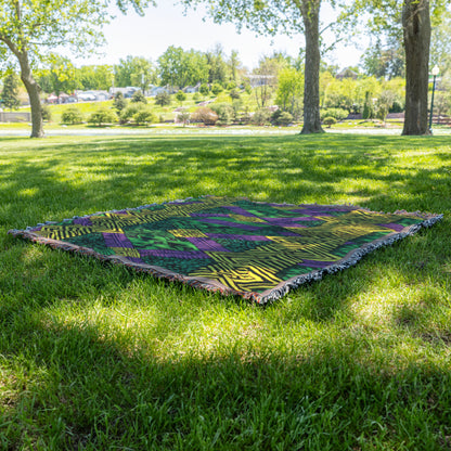 A vibrant throw blanket with green, purple, and yellow geometric patterns lies on green grass in a sunlit park. Trees cast dappled shade, with more greenery and houses visible in the background under a clear sky.