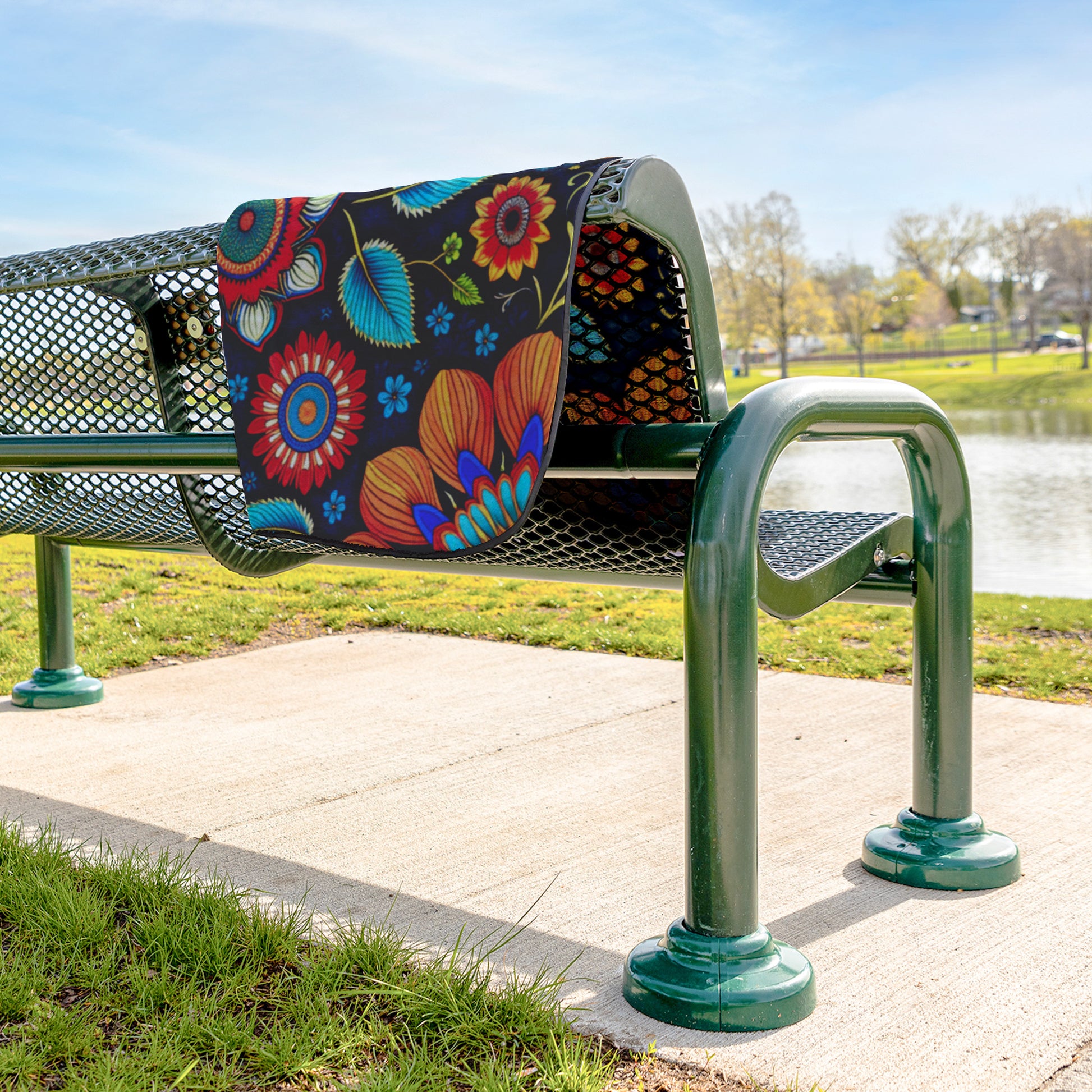 A metal park bench with a black seat and backrest has a plush fleece top stadium blanket draped over it. The blanket features bold, bright African-inspired patterns. The bench sits on a concrete path beside grass, a pond, trees, and blue sky.