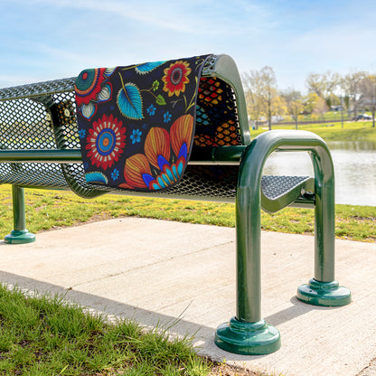 A metal park bench with a black seat and backrest has a plush fleece top stadium blanket draped over it. The blanket features bold, bright African-inspired patterns. The bench sits on a concrete path beside grass, a pond, trees, and blue sky.